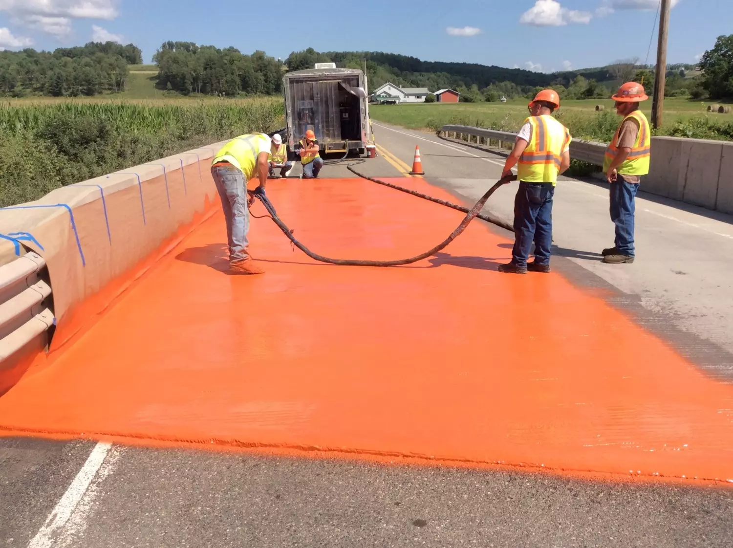An image of three construction workers using a sprayer to apply an orange coating to a bridge deck while two construction workers inspect the coating in the background.
